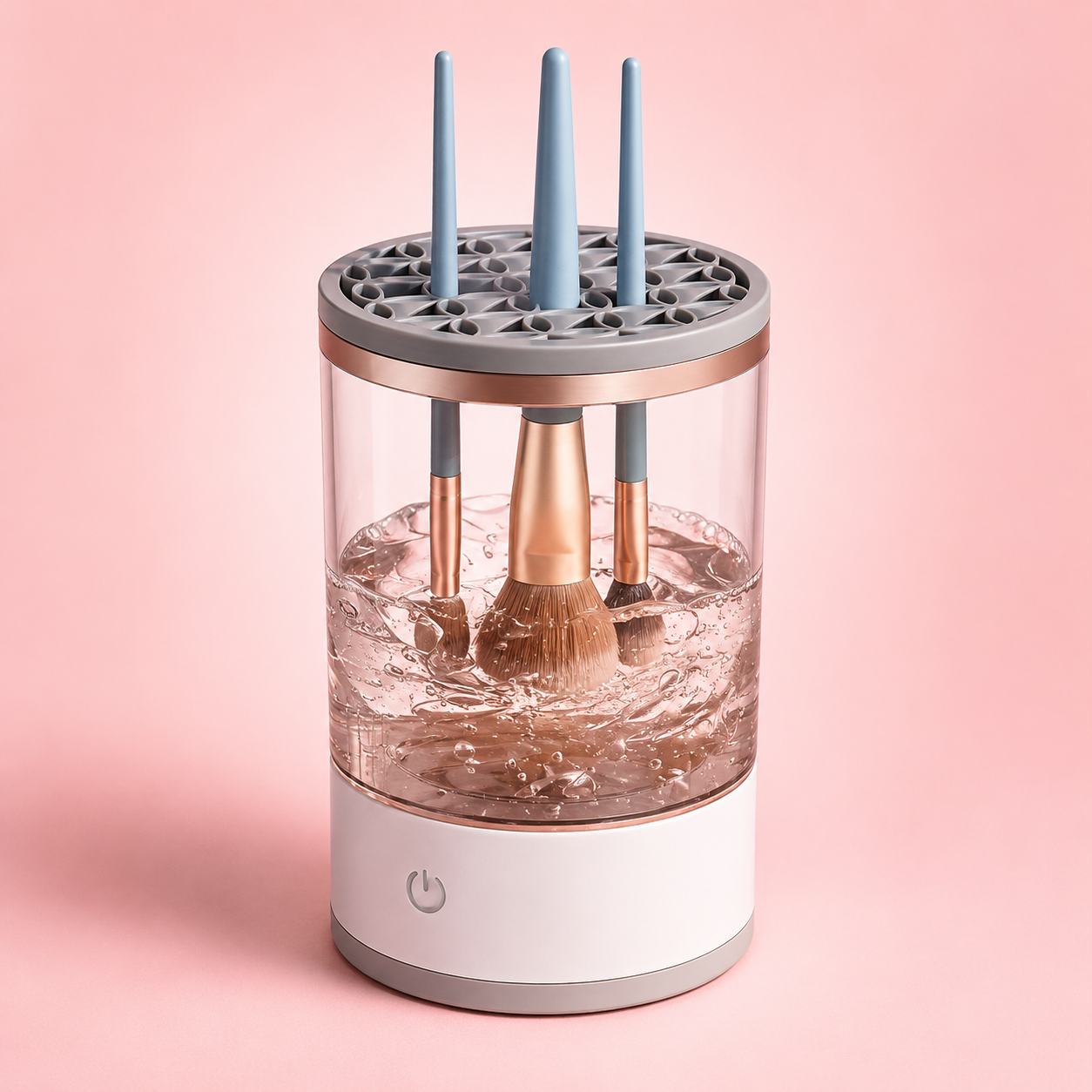 Automatic makeup brush cleaner and dryer with rose gold accents cleaning three makeup brushes in a clear water-filled chamber on a soft pink background.